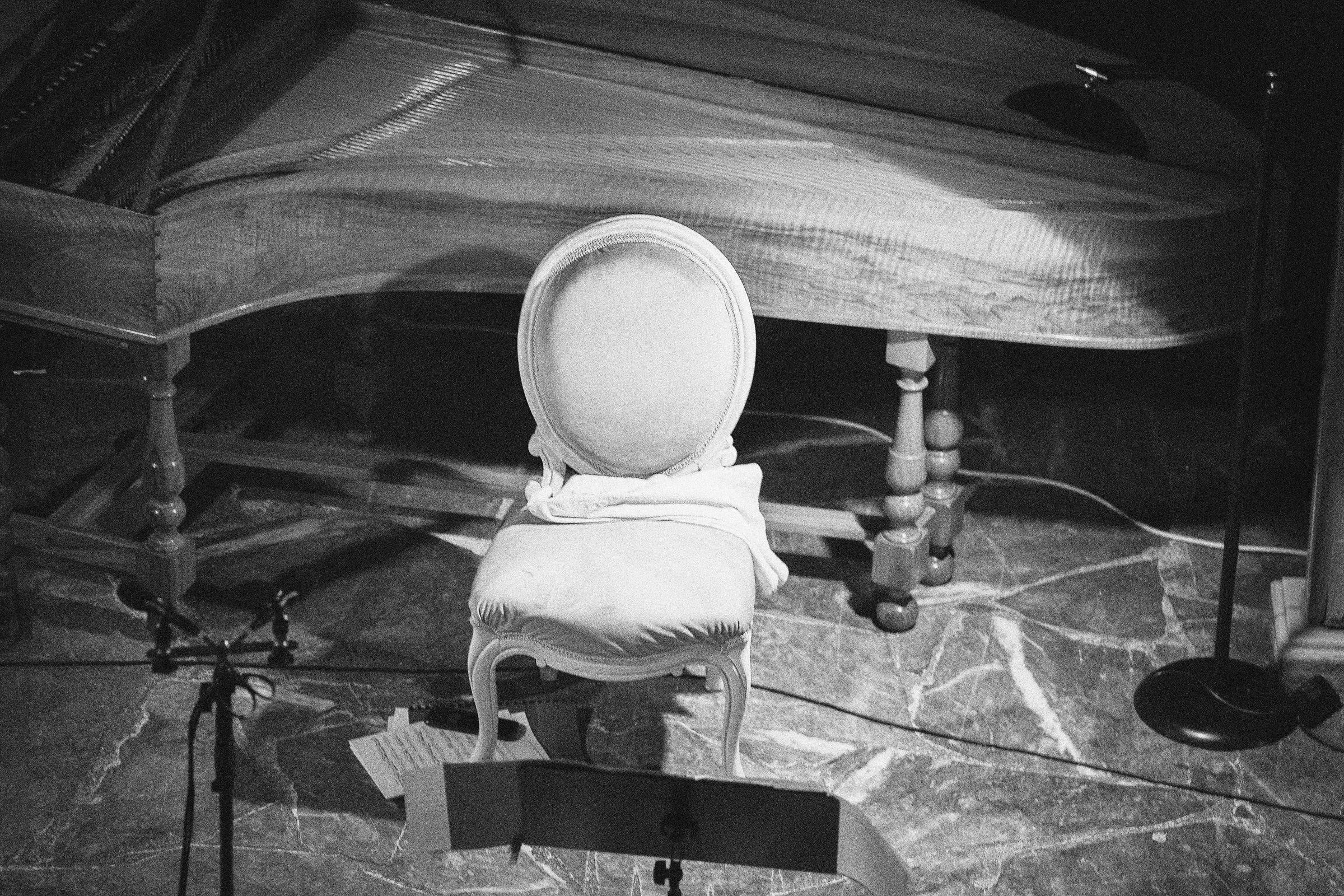 An empty upholstered chair and a music stand beside the harpsichord, on a marble floor.
