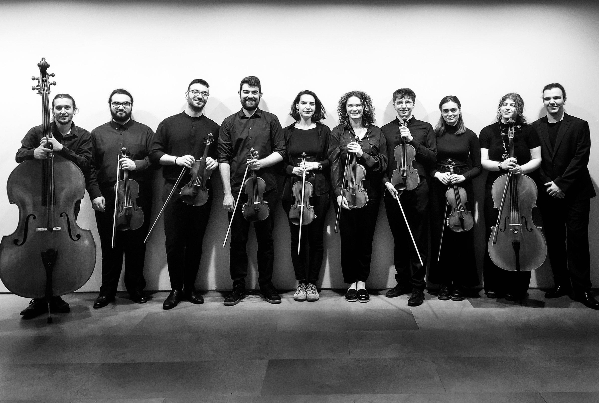 Ten musicians standing in a row against a white wall, each holding a string instrument.