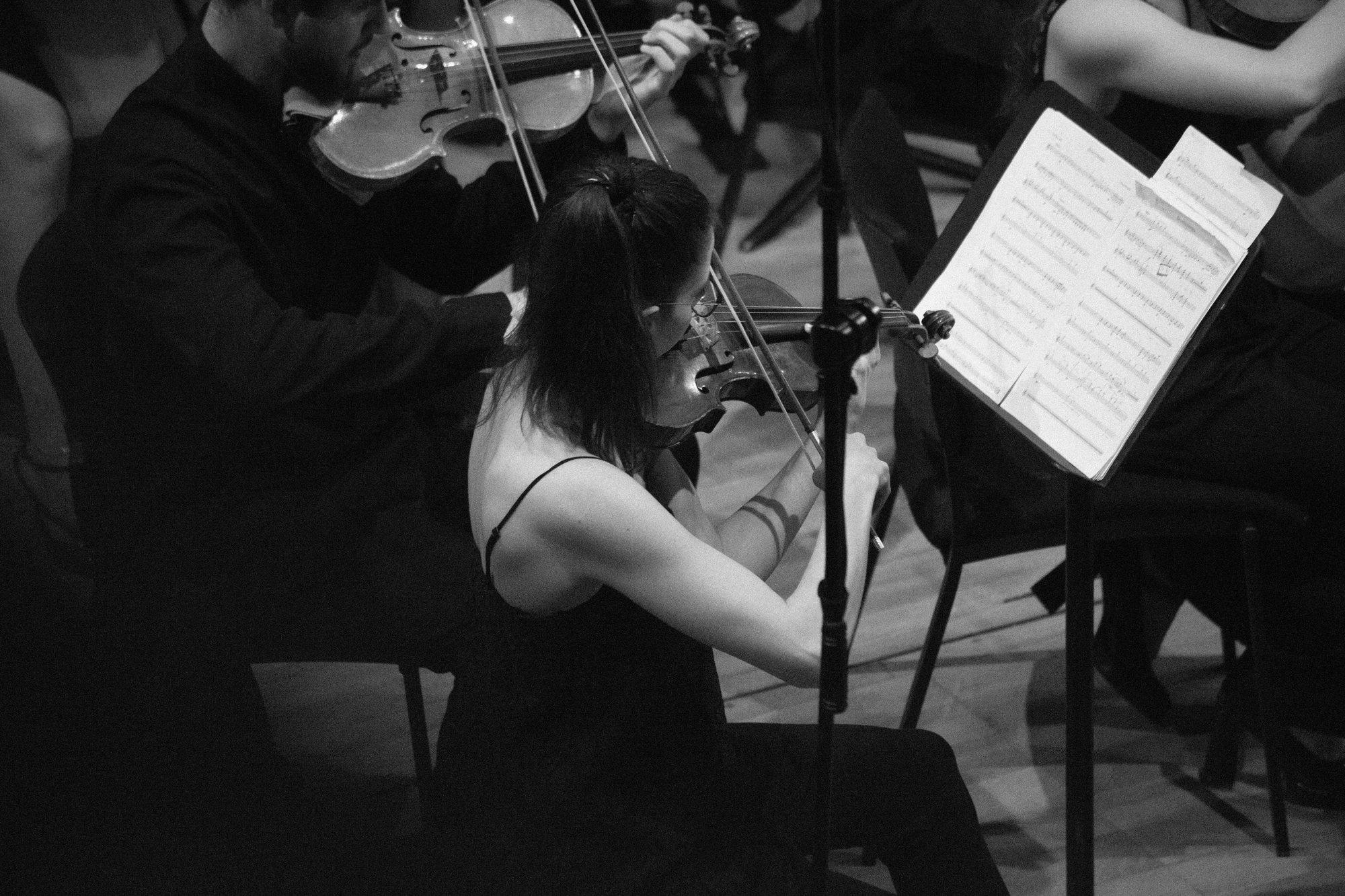 A violinist reading from a part on the stand, other string players close around her.