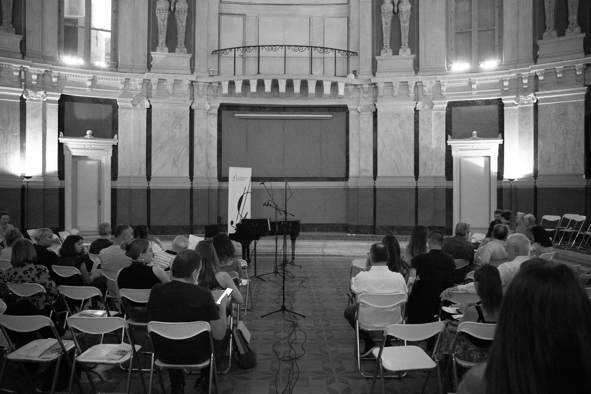 Le public, sur des chaises pliantes, dans une salle de marbre à galeries, face à un piano à queue et à des pupitres.