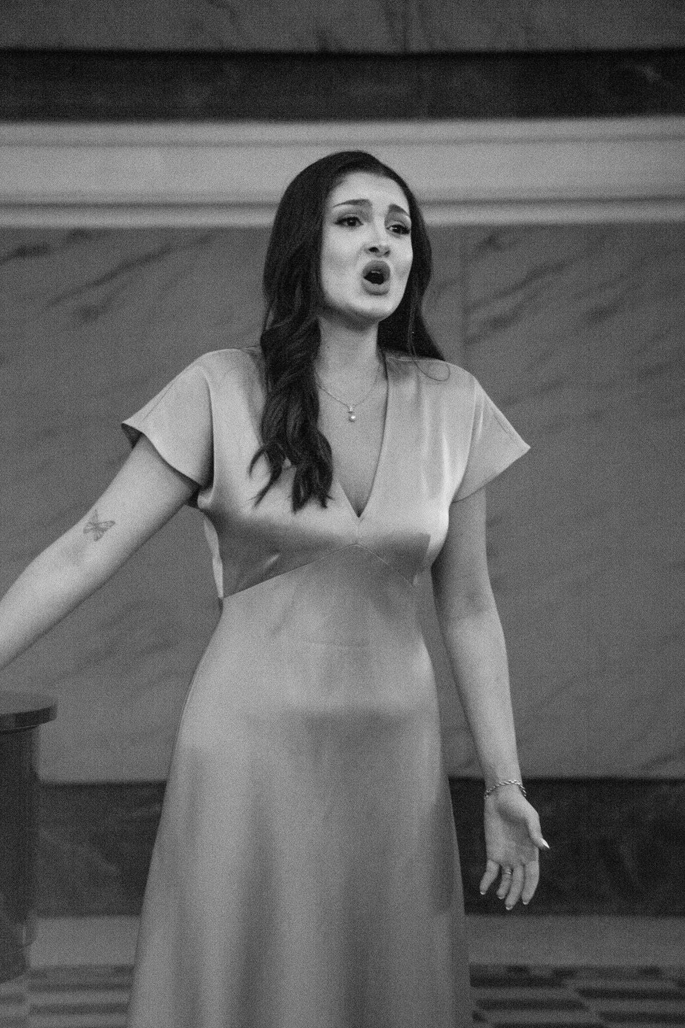 A singer in a long pale dress, mid-phrase, one hand open towards the audience.