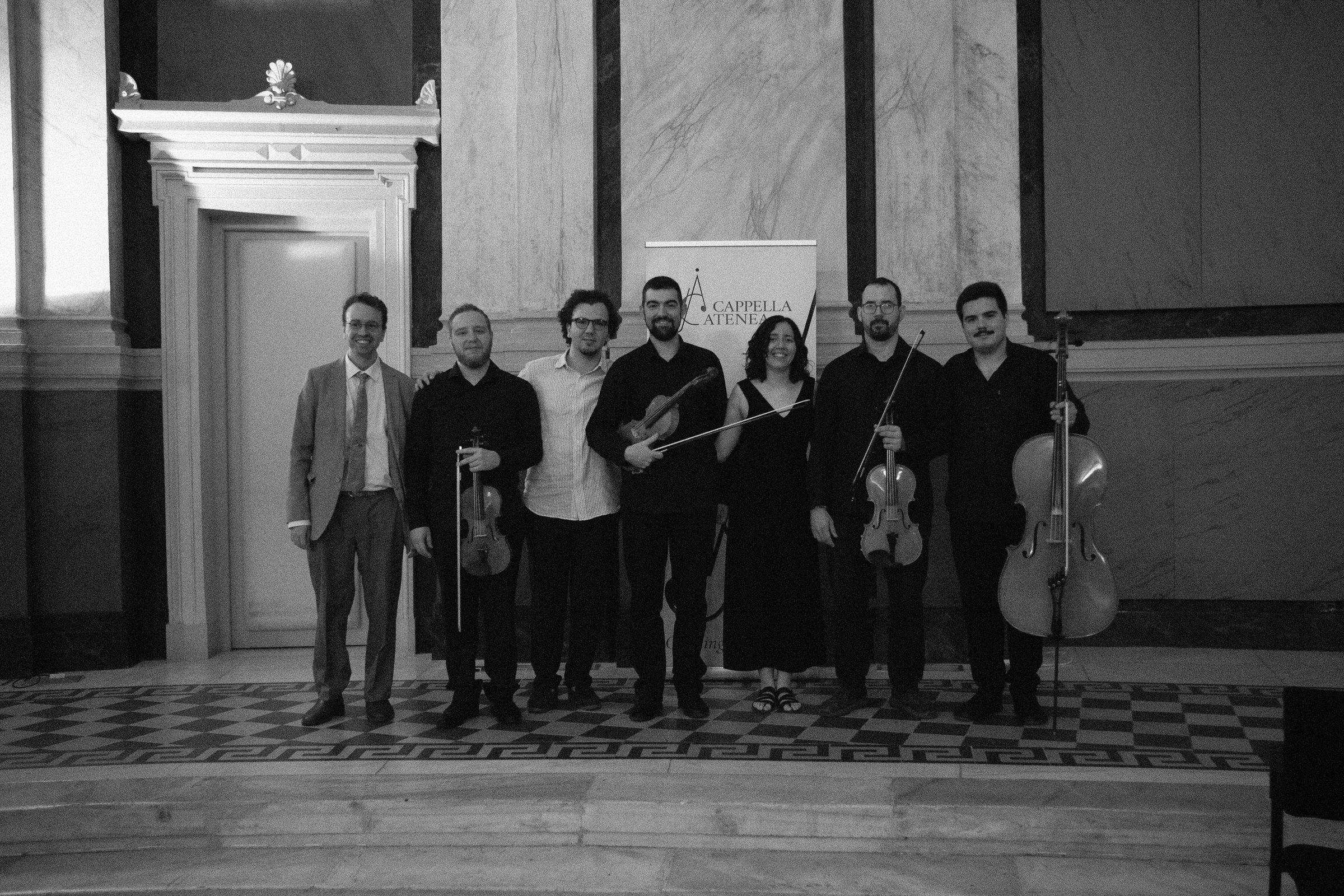 Sept musiciens debout en ligne avec leurs instruments sur un sol de marbre à motifs, saluant sous les applaudissements.