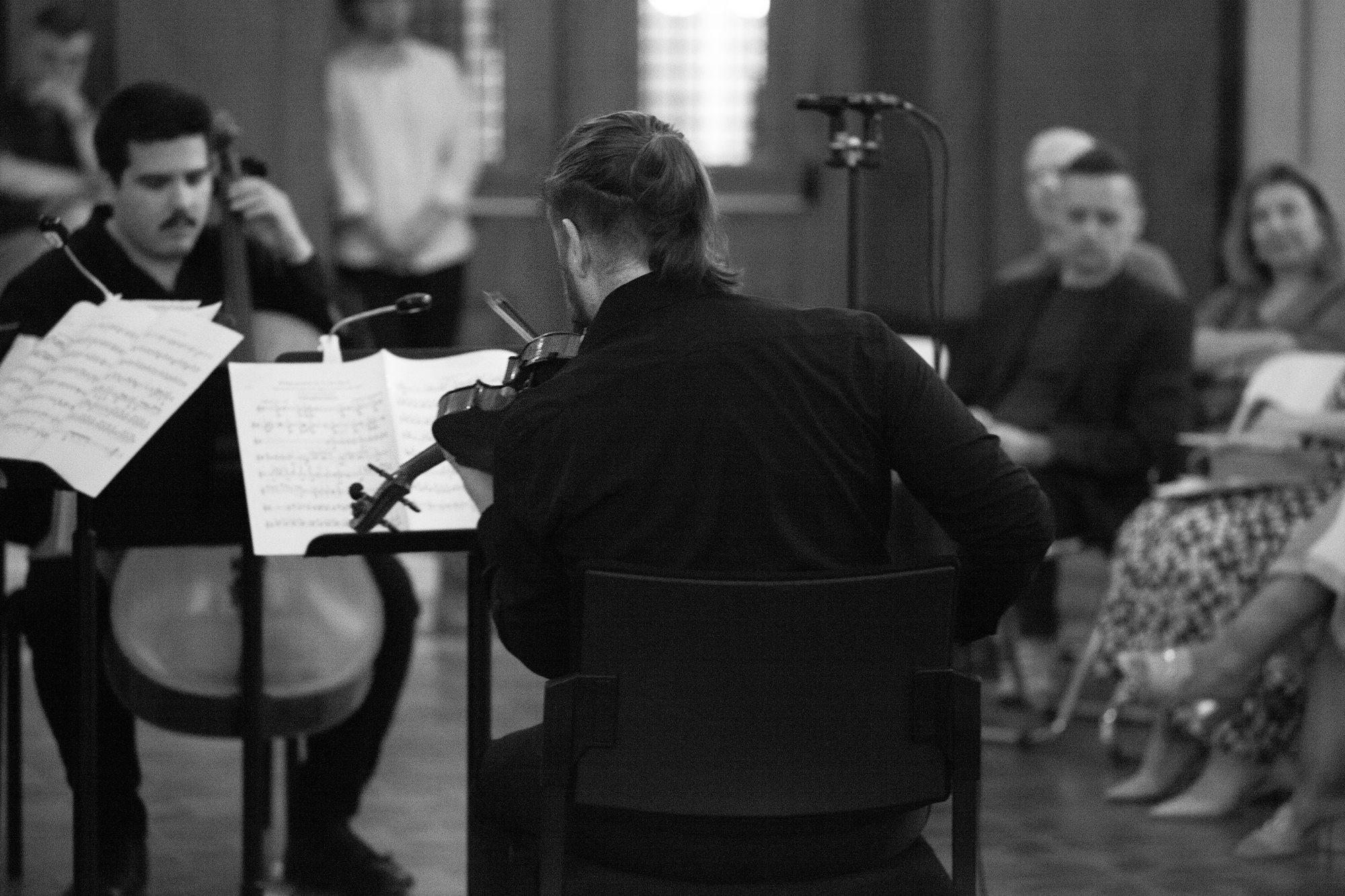 A cellist and a violinist play from shared stands, listeners seated close beside them.