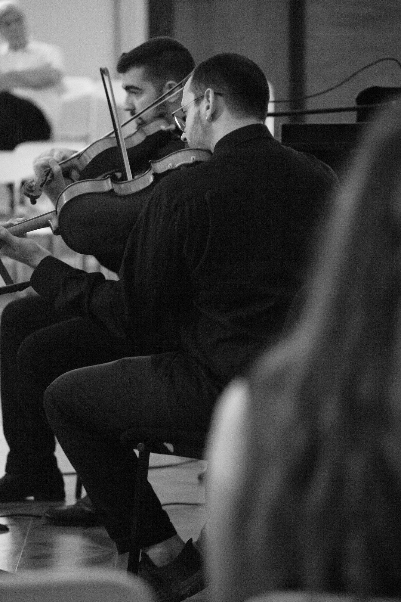 Two violinists playing side by side, seen past a listener’s shoulder.