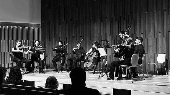 A string ensemble performing on a wooden stage in front of a panelled wall, the audience in silhouette.